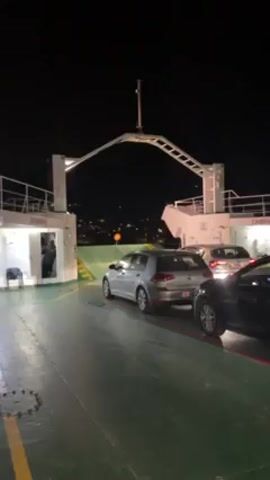 The video shows a car ferry at night. The ferry is filled with cars in neat rows, and a man is standing by a doorway. The ferry pulls away from the shore, and the lights of the land become visible across the water. Then another ferry is seen approaching in the distance.