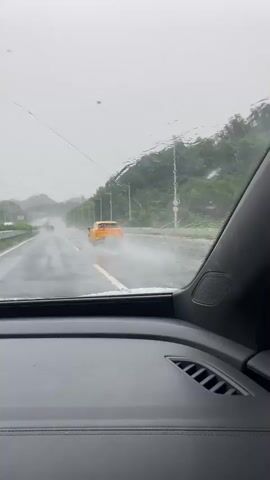 The video shows a view from inside a car on a highway during a rainstorm. The windshield is wet, with water streaks and some dirt. The road ahead is wet and appears to have standing water in some areas. Other vehicles are visible on the road. One car, a bright orange sports car, is driving in front of the camera car. The weather appears overcast, and there are trees and hills on the right side of the road. There are utility poles and streetlights lining the highway.