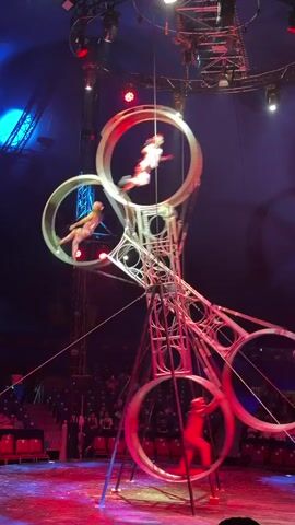 A circus performance is underway within a tent. The main attraction is a large, complex, metallic apparatus with four connected circular frames that rotate. Within these frames, three acrobats, dressed in red and white costumes, perform. One acrobat is suspended in mid-air within the topmost frame, seemingly defying gravity. Another acrobat is captured in motion within the frame to the right. The third acrobat is actively running inside the bottom left frame. The stage beneath them is illuminated with red and blue lights, casting dramatic shadows. The background shows the dark interior of the circus tent and audience seating in the distance.