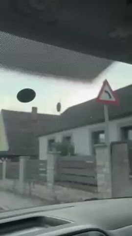 A camera inside a car looks out through the windshield as the car moves down a residential street. Houses are seen on either side, mostly single-family homes with fences or hedges. The sky is overcast.