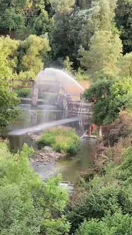 A body of water, likely a river or stream, flows between lush green trees. A concrete structure, possibly a dam or bridge, spans the water. A large water spray is being directed from the right side of the frame towards the left, creating a visible mist. There are multiple people and what appear to be machines, including an orange vehicle, on the right bank near the concrete structure. The scene appears to be an outdoor, natural environment with man-made elements.