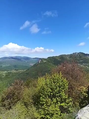 The video shows a scenic landscape on a sunny day. The foreground is characterized by lush green trees and rocky outcrops. In the distance, a small village is nestled amidst a verdant valley, surrounded by rolling hills and mountains. The sky is a clear blue, dotted with fluffy white clouds. The camera slowly pans across the landscape, offering a wide view of the natural beauty.