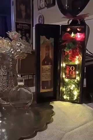 The video pans across a tabletop display. On the left is a decorative vase with iridescent flowers and a bottle of Chanel Chance perfume. To the right of the vase are two open wooden boxes, each containing a wine bottle. The left box has a rectangular image of a wine bottle on a distressed background. The right box contains a green wine bottle with a "ROJO" label and red grapes, illuminated by string lights. Red ribbon and green leaves are tied around the neck of the bottle. The background shows framed pictures on a wall, including a poster of The Bee Gees. The lighting is dim and warm, suggesting an indoor setting, possibly a home decor arrangement.