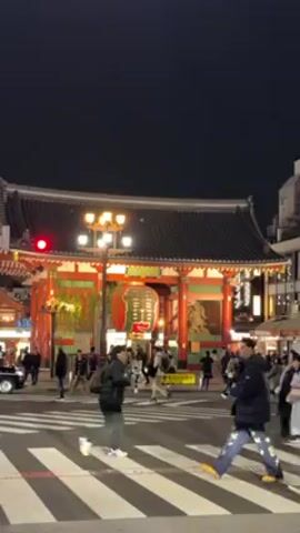 The video depicts the Kaminarimon gate in Tokyo, Japan at night. The gate is brightly lit, with a large red lantern hanging in the center. People are walking across a crosswalk in front of the gate, and there are shops visible on either side of the street. The sky is dark, and the overall scene is busy with activity.
