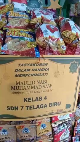 The video shows a display of snacks and treats, likely for an event. A sign in the center reads "TASYSURAN DALAM RANGKA MEMPERINGATI MAULID NABI MUHAMMAD SAW," which translates to "Thanksgiving in Commemoration of the Prophet Muhammad SAW's Birthday." Below this, it says "KELAS 4 SDN 7 TELAGA BIRU," indicating it's for 4th grade at SDN 7 Telaga Biru. The snacks are arranged in bags and boxes, with some stacked high. In the background, there are decorations like stars and a crescent moon. A person, possibly an adult, wearing a headscarf, is visible in the lower left corner, partially obscuring the view. The environment appears to be indoors, possibly a school or community hall.