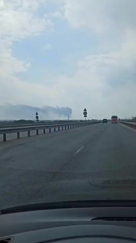 A car is driving on a highway. In the distance, a large plume of dark smoke rises into the sky. There are other vehicles on the road. The sky is partly cloudy. Road signs are visible on the side of the highway. A railing is on the side of the highway.