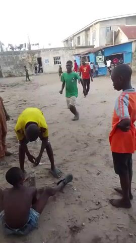 The video shows an outdoor scene in what appears to be a sandy or dirt-filled yard. Several children are present, with one lying on his back on the ground. Another child, wearing a yellow and black shirt, is standing next to him. Two other children are standing nearby. There are buildings in the background, some with laundry hanging on the lines. Debris is scattered around the ground.