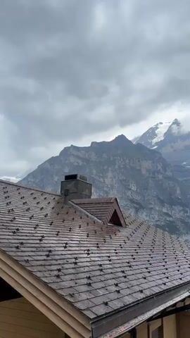 The video shows a close-up view of a dark, sloped roof with shingles. Behind the roof, a majestic mountain range with snow-capped peaks is visible under a cloudy sky. The mountains appear rugged and steep, with some vegetation clinging to their slopes. The overall scene suggests a location in a mountainous region, possibly a village or resort, with a focus on the natural landscape.