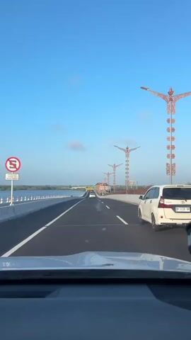 The video shows a view from the driver's seat of a vehicle traveling on a bridge. The bridge has multiple lanes of traffic and is lined with decorative lampposts featuring stylized figures. On the left side of the bridge, there is a body of water and a white barrier. Several cars are visible on the bridge, including a white van and a black SUV. The sky is clear and blue, with a few wispy clouds. The overall environment appears to be a highway overpass, possibly in a coastal or riverside area, under daylight conditions.