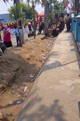 The video shows a street scene with a group of people, some in uniform, seemingly preparing for an event. There are palm trees, a drainage ditch, and some trash visible. In the background, there are flags and a red tent. The people are standing and sitting along the side of the road, and some appear to be children in uniform. The overall atmosphere suggests a community gathering or a parade.