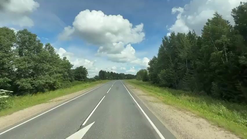 The video captures a first-person perspective of driving on a paved, two-lane highway with white lines marking the lanes. The road is flanked by lush green grass and dense trees on both sides, indicating a rural or natural setting. The sky is a vibrant blue, dotted with large, fluffy white clouds, suggesting fair weather. The road appears to be relatively straight for the most part, with gentle curves. There are occasional power lines and poles visible along the roadside. Traffic signs are also present, including what appear to be circular signs with a red border and a line through them, indicating a prohibition. The overall impression is of a peaceful drive through a scenic landscape on a clear day.