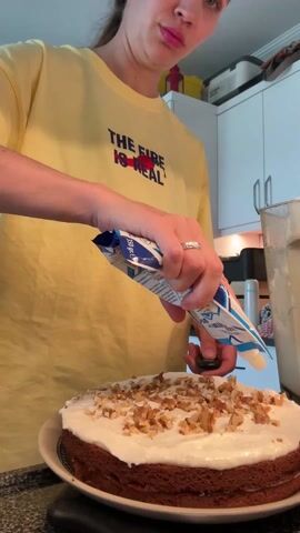 In a brightly lit kitchen, a woman wearing a yellow t-shirt with the words "THE FIRE IS REAL" is decorating a cake. She is squeezing a blue and white package, letting a white liquid drizzle onto a round cake. The cake is topped with a layer of white frosting and scattered chopped walnuts. A blender containing a white substance is visible to the right of the cake. The kitchen features white cabinets and various other kitchen items in the background.