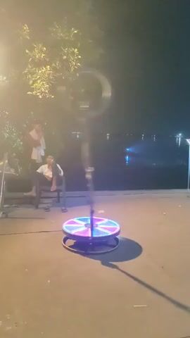 This video shows a 360-degree photo booth set up outdoors at night. The booth has a circular platform with colored LED lights that rotate. A ring light is attached to a stand behind the platform. Two individuals are visible in the background, one standing and one sitting on a chair. Trees and water are also present in the background. The ground appears to be concrete.