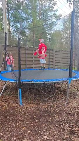 A boy wearing a red hockey jersey with the number 91, a red baseball cap, and plaid shorts is jumping on a trampoline outdoors. A girl wearing a pink t-shirt and blue jeans is standing to the left of the trampoline, near the ladder. The trampoline has a blue safety net and a blue edge. The ground around the trampoline is covered in mulch and some dry leaves. A wooden fence is visible in the background, along with trees.