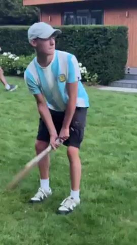 A young man wearing a striped Argentina soccer jersey, black shorts, white socks, and athletic shoes swings a cricket bat on a green lawn, then raises his hand to his left while looking in that direction. A second person in a white t-shirt and red shorts stands in the background. Behind the first man are a green hedge and a small building with dark wood siding.
