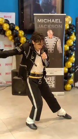 A man dressed as Michael Jackson performs in front of a banner featuring the singer and a backdrop of gold and black balloons. He is wearing a black and white costume with a white shirt, black pants with white stripes, and black shoes. He is holding a microphone and appears to be singing and dancing. The environment appears to be an indoor event space with blue walls and a wooden floor.