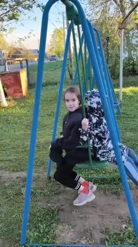 Two children are on a playground swing set. The swing set is made of blue and green metal. A girl with long brown hair in a black top and black pants with pink sneakers sits on one swing, while a child wearing a black and white camo outfit with a hood up sits behind her. They are both swinging. The ground is grass and dirt, with a small brown shed visible in the background. A car is visible driving down a road in the distance. The sky is clear and the lighting indicates it is daytime.