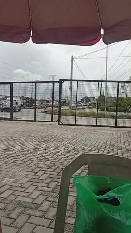 The video is a first-person perspective shot from under a burgundy umbrella. In the foreground, a gray plastic chair is visible, with a bright green plastic bag containing dark items resting on it. The background shows a paved area with interlocking brick pavers. Beyond that is a large black metal gate. Through the gate, a street scene is visible with various cars and a white bus. The sky is overcast. The gate appears to be opening or closing at the beginning of the video. A grey sports car is seen passing through the gate at the beginning of the video.