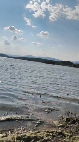 This media file shows a scenic view of a body of water, likely a lake or reservoir, with a shore in the foreground. The water is rippled, suggesting a light breeze. The sky is blue with scattered white clouds, indicating a pleasant daytime weather condition. In the distance, rolling hills or mountains form a natural backdrop, and a small structure, possibly a building or a boat, is visible on the water or near the far shore. The foreground of the image depicts the edge of the water, with some vegetation and debris on the sandy or muddy shore. The overall impression is one of a serene, natural landscape, possibly enjoyed during a calm day. There is no visible text in this media file. The location is identified as Orizari, Bulgaria.