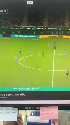 The video displays a computer monitor showing a soccer match in progress. The screen shows a live broadcast with game statistics, player positions on the green field, and audience in the stands. A chat window is visible on the right side of the monitor with user comments. The lower part of the monitor displays a small, blue USB fan placed in front of it. Several sticky notes with handwritten text are affixed to the monitor's bezel.