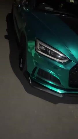 The video captures the front and right side of a car parked outdoors at night. The car features a distinctive iridescent or chameleon-like wrap, primarily appearing as a vibrant metallic green under the low light, with hints of blue in its reflections. It is equipped with multi-spoke black wheels, through which red brake calipers are visible. The side mirror on the driver's side is covered in a carbon fiber-like wrap. A black front splitter is also visible beneath the grille.

The camera pans slowly from the car's front right corner, moving along its right side towards the rear. The scene is dimly lit, causing the car's reflective surface to stand out against the dark asphalt or concrete ground. The overall setting appears to be a parking area or street, consistent with the South Kesteven, United Kingdom context provided. No specific landmarks are visible. An audible laugh can be heard in the background, suggesting the presence of people, though none are seen in the frame. There is no visible text. The time of day is clearly night.