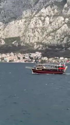 The video shows a maroon tour boat moving on the water with a mountain and city visible in the background. The boat has two levels and is carrying passengers. The mountain is light gray with some green vegetation. The city has white buildings with red roofs. The water is dark blue and choppy. There is some wind noise evident in the audio.