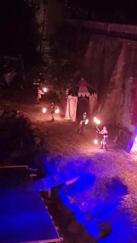 The media file depicts an outdoor performance at night, possibly a festival or a theatrical event. The scene is set in a rustic environment with a large stone wall as a backdrop, illuminated by purple and blue lights. A small, tent-like structure with a red and white striped canopy is visible on the left, suggesting a medieval or fantasy theme.

Several individuals, dressed in what appear to be historical or costume-like attire, are engaged in a performance. They are interacting with fire, holding lit torches or performing with flames. Some individuals are moving around, while others appear to be in the process of manipulating fire. The ground is covered in dirt or sand, and there are some wooden structures or platforms in the foreground, also illuminated by blue light. A long red fabric hangs vertically from the top of the stone wall, adding a splash of color to the scene.

The overall atmosphere is dramatic and energetic, with the flickering firelight creating a sense of mystery and excitement. The time of day is clearly night, and the lighting suggests an evening event. The location, Sant Joan Les Fonts, Spain, hints at a possibly historical or cultural celebration. No visible text is present in the media file.