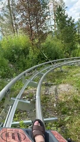 POV shot from a gravity coaster as it travels along metal tracks. The coaster is surrounded by green trees and vegetation. The sky is visible in the background with some clouds. The rider's feet are visible in the foreground, wearing sandals. The track curves and goes uphill.