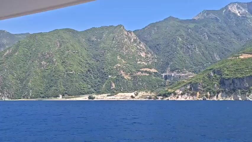A wide shot captures a mountainous coastline with lush green vegetation meeting a deep blue sea. The view is from a boat, with a sliver of its white structure visible at the top of the frame. The terrain is rugged and steep, with some rocky outcrops and a winding road visible on the mountainside. A cluster of buildings, possibly a monastery or settlement, is perched on a plateau in the middle distance, blending into the landscape. The sky is clear and blue. The video appears to be a steady shot, possibly taken while traveling along the coast.