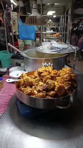 The video shows a busy food stall in what appears to be a market or food court setting. A large, silver pan filled with golden-brown cooked food sits prominently in the foreground on a scale, near a large pot and other dishes. The camera then pans to show a row of simple wooden tables where people are seated, eating. Many other food stalls are visible in the background, with people walking around and purchasing food. The area is covered by a roof, possibly a temporary structure. People are casually dressed, and the overall impression is of a lively, crowded, and informal dining area.