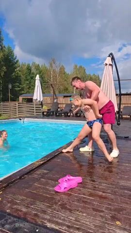 At 00:00, a man in red shorts is pushing a boy in blue patterned swim trunks towards the edge of a swimming pool. A woman is in the pool. At 00:01, the boy jumps into the pool. At 00:02, the boy lands in the pool, creating a splash. The man watches from the poolside. At 00:03, the boy and woman are swimming in the pool. At 00:04, the boy and woman are swimming. At 00:05, the boy and woman are swimming. The environment is outdoors with a swimming pool, wooden deck, lounge chairs, umbrellas, and trees in the background. There is a pair of pink flip-flops on the deck.