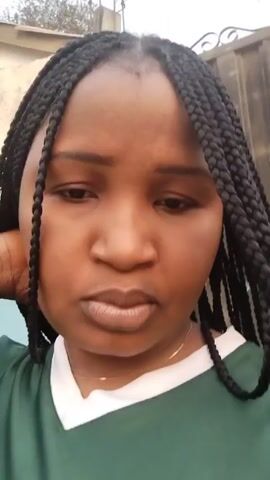 A close-up shot captures a person with medium brown skin and braided black hair, looking directly at the camera. The individual wears a green shirt with white trim. The background is slightly blurred, revealing a building or wall with muted colors. A vehicle horn is heard distinctly. The person touches their hair and looks into the camera.