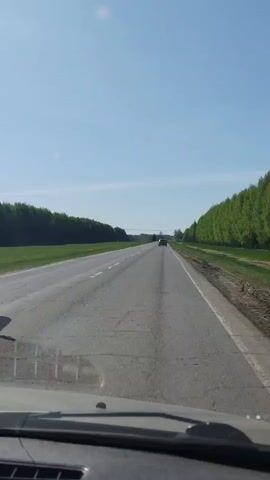 The video, shot from the interior of a car, shows a stretch of gray asphalt highway under a bright blue sky. The road is flanked by green trees on either side, forming a corridor-like perspective. Another vehicle, a dark-colored SUV, is seen driving ahead in the same direction, with another car in the far distance. The camera captures the view through the windshield, including a section of the dashboard. The road surface appears cracked in places, indicating wear. The overall impression is a rural road trip on a clear day.