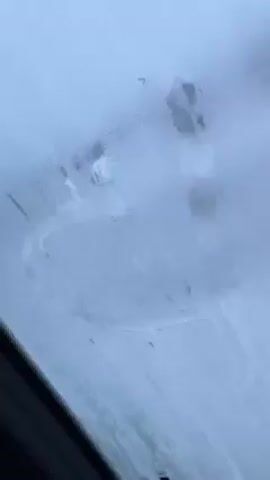 The video shows a car windshield that is fogged up. A circle has been partially wiped away, revealing the cloudy view outside. Small water droplets cover the remaining areas of the glass. The car's interior frame is visible along the bottom left edge of the shot. The camera angle is from inside the car, looking upwards through the windshield.