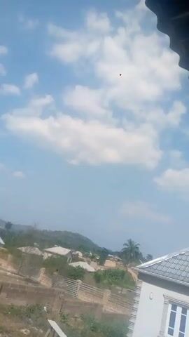 The video shows a daytime outdoor scene with a view of a neighborhood under a blue sky with scattered white clouds. In the foreground are rooftops of buildings, a brick wall, and some greenery. In the background are more houses and a distant hillside. There are sounds of traffic and possibly a siren. The camera seems to be held from a slightly elevated position.