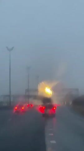 The video shows a POV shot of driving on a highway in heavy fog and rain. The road is wet and appears to have white lane markings. There are streetlights visible along the side of the road and overhead structures, possibly overpasses or signs. Several cars are ahead, with their brake lights illuminated, indicating reduced visibility and traffic. The overall atmosphere is dark and hazy, suggesting poor weather conditions. There are no people clearly visible, and the focus is on the driving environment.