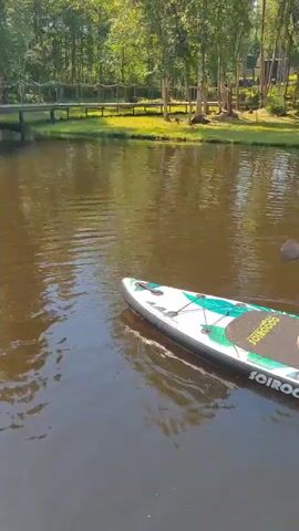 A person is paddleboarding on a calm body of water. The person is kneeling on the paddleboard, holding a paddle and appears to be paddling forward. The paddleboard is white with blue and green accents and has the brand name "SCIROCCO" printed on it. The water is dark and reflects the surrounding trees. The background shows a lush green forest with birch trees and some structures visible on a hill. The sky appears clear, suggesting a sunny day. The overall scene is peaceful and suggests a recreational activity. The time of day appears to be daytime, possibly late morning or afternoon, given the lighting. The location is Zashchita, Russia.