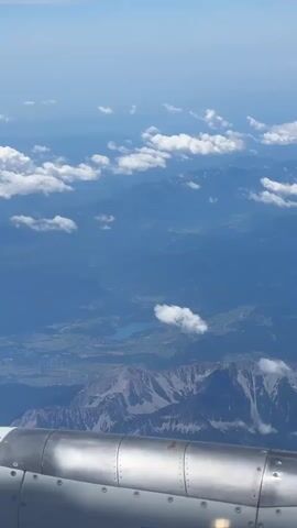 This is a vertical, aerial view from an airplane window. The foreground features the metallic engine housing of the aircraft, which is silver and cylindrical with visible rivets and seams. The background displays a vast landscape of mountains and valleys under a clear blue sky dotted with white cumulus clouds. A bright blue lake is visible nestled within a valley between mountain ranges. The overall impression is of flight over a scenic, mountainous region.