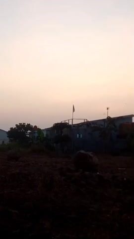 The video shows an outdoor scene, likely during dawn or dusk, as indicated by the soft, diffused light. The foreground is dominated by dark, tilled soil, suggesting a construction site or agricultural land. In the mid-ground, there are some scattered rocks and vegetation. Behind the soil, several structures are visible. To the left, a large, light-colored building with a sloped roof can be seen. In the center, a smaller building with a flag on top is present. Further to the right, another building with lights on its facade is visible. Trees are scattered throughout the scene, with some appearing denser on the left side. The sky is a pale, hazy color. There are no people visible in the video. The overall impression is one of a developing or industrial area at the edge of a natural landscape.