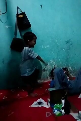 The video shows a young child in a striped t-shirt and dark shorts standing in a room with light blue walls. The child is smiling and appears to be dancing or moving energetically. On the floor, which is covered with a red patterned rug, there are several objects including a dark-colored bag hanging on the wall, a box, and some green and white items that appear to be toys. In the background, another person is lying down and looking at a phone. The room seems to be dimly lit.