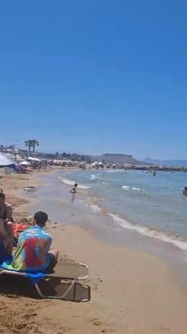 A daytime video shows a sandy beach under a clear blue sky. Gentle waves break on the shore of a clear, blue-green sea where several people are swimming and wading. A rocky breakwater is visible further out in the water. In the foreground, a person wearing a colorful shirt is seated on a beach chair, partially visible on the left. Later in the video, a small child in a light-colored swimsuit is seen playing in the shallow water with a pink bucket and a red shovel. Distant mountains line the horizon.