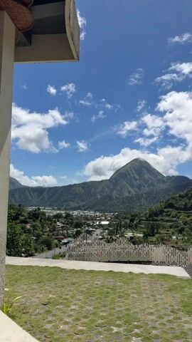The video shows a beautiful scenic view of a lush, green mountain, likely a volcano, partially shrouded in clouds against a bright blue sky dotted with fluffy white clouds. At the base of the mountain sits a small town nestled among the hills and greenery. The foreground features a patterned grass lawn and a white fence, with a glimpse of a building’s structure in the frame's corner, adding perspective. The camera zooms in gradually on the mountain, providing a closer look at its terrain and vegetation. The scene is peaceful and vibrant, showcasing nature's beauty on a clear, sunny day.