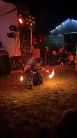 A person is performing fire dancing in an outdoor area at night. The performer is wearing a dark top and a patterned skirt, and is holding two flaming torches. The area is dimly lit with stage lights and string lights visible in the background. There are a few other people in the background watching the performance. The ground is covered in grass and dry leaves.