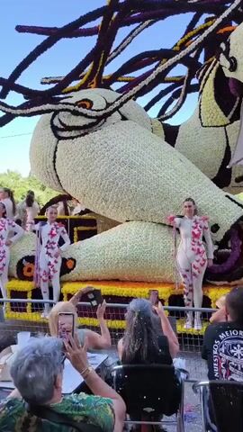 The video shows a parade float decorated with white flowers, resembling a large creature with a head and long, dark tendrils. Several women in matching white costumes with butterfly patterns are posing on the float. In the foreground, spectators are seated, many of them taking photos or videos of the parade with their phones. The scene appears to be outdoors during the day, with a clear blue sky visible.