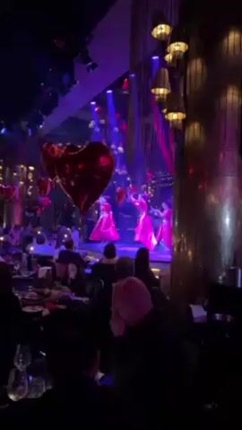 In a dimly lit restaurant, a stage is set up with three dancers in red outfits performing. They hold heart-shaped balloons and move in unison, illuminated by blue and red stage lights. Diners sit at tables, observing the performance. Heart-shaped balloons decorate the foreground. The interior is decorated with hanging lights and patterned columns.