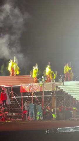 The video captures a nighttime outdoor event with multiple stages and performers. In the foreground, people stand in silhouette against a brightly lit stage with scaffolding and stairs. The stage features performers in yellow tops and red pants, some holding illuminated objects, and others appear to be interacting with pyrotechnics. Smoke and flares in red and yellow hues are visible, creating a dynamic and visually striking atmosphere. The background reveals a building with illuminated windows. The lighting is dramatic, with spotlights and bursts of light illuminating the performers and the stage. The overall impression is that of a lively performance or festival.