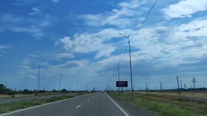A POV video captures a road trip with a blue sky filled with fluffy white clouds. Streetlights line the road, and power lines stretch across the horizon. A billboard with Russian text and a QR code is visible on the right. In the distance, a few cars are driving on the road, and power lines are scattered throughout the landscape. The road is asphalt and appears to be well-maintained. The surrounding landscape is dry and grassy. The video is shot from a moving vehicle, suggesting the perspective of a driver or passenger.