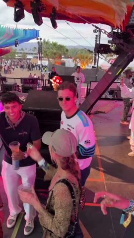 The video shows an outdoor music festival with a large crowd of people. The camera pans from a group of people on stage to show the audience in front of the stage. The stage is decorated with colorful flags and lights. There are speakers and other equipment on the stage. The audience is made up of people of all ages and backgrounds. They are all dressed casually and are enjoying the music.