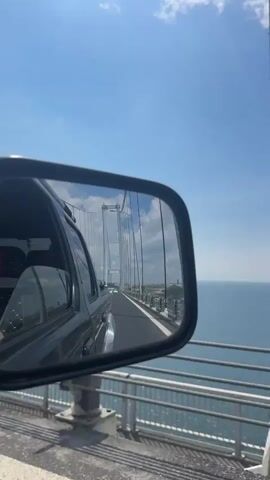 The video shows a view in a side mirror of a car traveling on a bridge. The bridge appears to be a suspension bridge, with large cables visible in the reflection. The sky is blue with scattered clouds, and the water is a light blue. The car in which the video is filmed is dark, and the side mirror is black. The bridge has gray railings on either side of the road. There are other cars on the bridge.