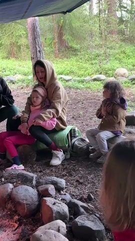 A group of people are sitting around a campfire under a tarp in a wooded area. In the center, a woman wearing a beige hooded sweatshirt is holding a young girl in her lap. The girl is wearing a pink shirt and a pink headband. To the right, a child with curly hair wearing a brown jacket with a purple hood is sitting with their hands clasped to their chest. In the foreground, more children are visible from behind, facing the fire. The ground is covered in dirt and rocks, and the surrounding area is filled with trees and foliage.